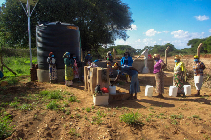 A queue of women wait at a water point in front of a water tank and solar panel set up